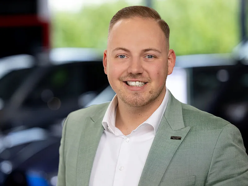 Kaltenbach Gruppe – Ein lächelnder Mann mit kurzen blonden Haaren, der einen hellgrünen Blazer und ein weißes Hemd trägt, steht in einem Haus in Kaltenbach Bergisch Gladbach, mit unscharfen Autos und Grünflächen im Hintergrund. – BMW, MINI, KIA, BMW Motorrad & Honda Motorrad Partner in NRW