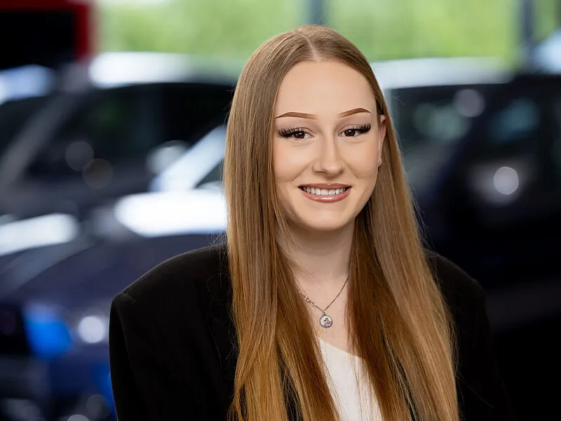 Kaltenbach Gruppe – Eine junge Frau mit langen glatten blonden Haaren, die einen schwarzen Blazer und ein weißes Oberteil trägt, lächelt in die Kamera. Im Hintergrund sind unscharfe Autos und Grünanlagen in Bergisch Gladbach zu sehen, die auf die lebendige Umgebung Kaltenbachs hinweisen. – BMW, MINI, KIA, BMW Motorrad & Honda Motorrad Partner in NRW
