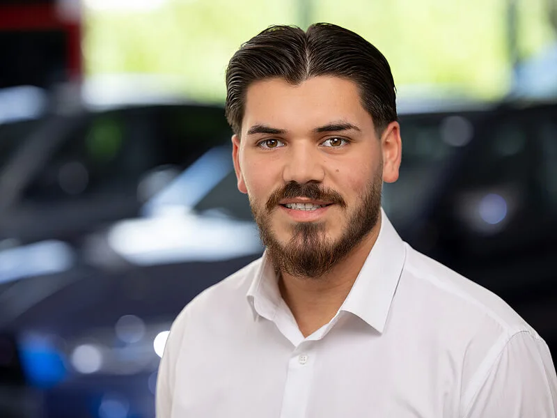 Kaltenbach Gruppe – Ein junger Mann mit dunklen Haaren und einem Bart, der ein weißes Hemd trägt, lächelt in die Kamera. Der Hintergrund ist unscharf und deutet auf Autos und Grünflächen hin, möglicherweise in der Nähe von Kaltenbach in Bergisch Gladbach. – BMW, MINI, KIA, BMW Motorrad & Honda Motorrad Partner in NRW