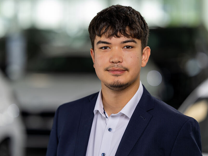 Kaltenbach Gruppe – Ein junger Mann mit kurzen dunklen Haaren und heller Gesichtsbehaarung, der eine marineblaue Anzugsjacke und ein weißes Hemd trägt, steht in einem Haus in Kaltenbach Lennestadt vor verschwommenen Autos. – BMW, MINI, KIA, BMW Motorrad & Honda Motorrad Partner in NRW