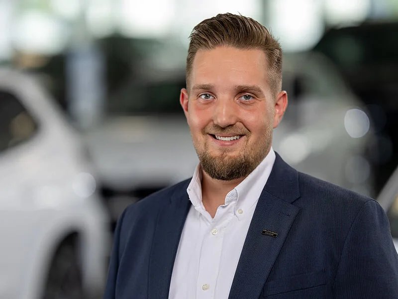 Kaltenbach Gruppe – Ein Mann mit kurzen, gestylten braunen Haaren und einem gestutzten Bart, der einen marineblauen Blazer und ein weißes Hemd trägt, lächelt in die Kamera. Die verschwommenen Autos und die hellen Lichter im Hintergrund deuten auf das Autohaus Kaltenbach Bergisch Gladbach hin. – BMW, MINI, KIA, BMW Motorrad & Honda Motorrad Partner in NRW