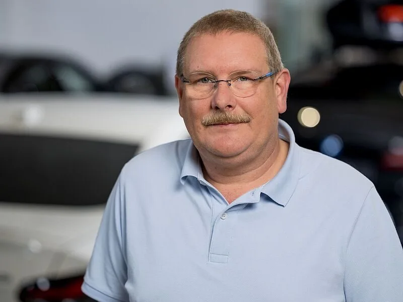 Kaltenbach Gruppe – Ein Mann mittleren Alters mit kurzen Haaren, Brille und Schnurrbart, der ein hellblaues Poloshirt trägt, steht in einem Innenraum vor verschwommenen Autos in Kaltenbach Lüdenscheid. – BMW, MINI, KIA, BMW Motorrad & Honda Motorrad Partner in NRW