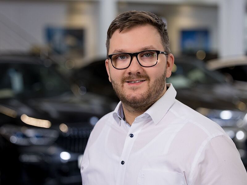 Kaltenbach Gruppe – Ein Mann mit kurzen Haaren und Bart, der eine Brille und ein weißes Hemd trägt, steht in einem Innenraum mit unscharfen Autos im Hintergrund, wahrscheinlich im Autohaus oder Autohandel Kaltenbach Lüdenscheid. – BMW, MINI, KIA, BMW Motorrad & Honda Motorrad Partner in NRW