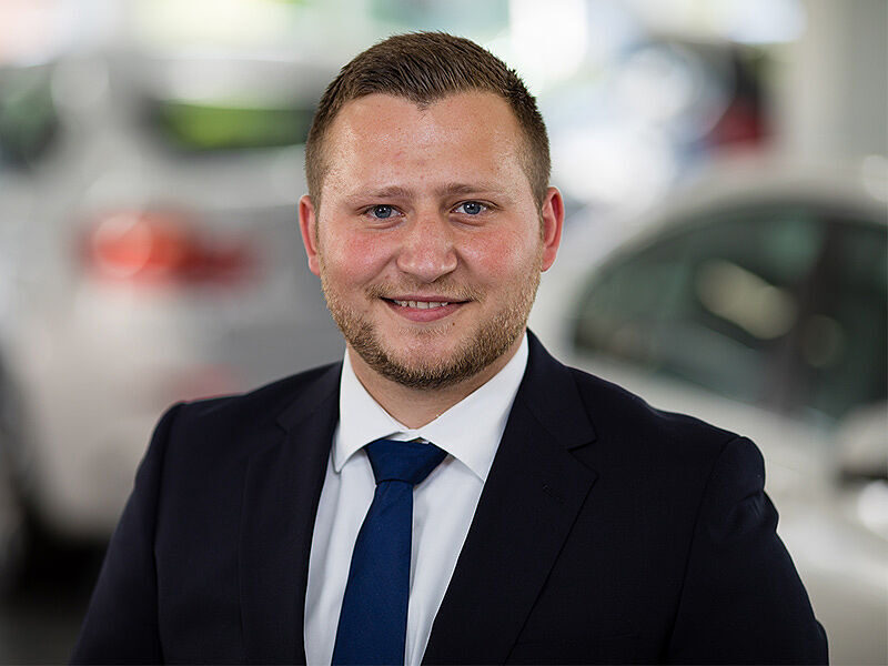 Kaltenbach Gruppe – Ein Mann im Anzug und mit blauer Krawatte lächelt in die Kamera. Er steht in einem scheinbaren Ausstellungsraum von Kaltenbach Remscheid mit unscharfen Fahrzeugen im Hintergrund. – BMW, MINI, KIA, BMW Motorrad & Honda Motorrad Partner in NRW