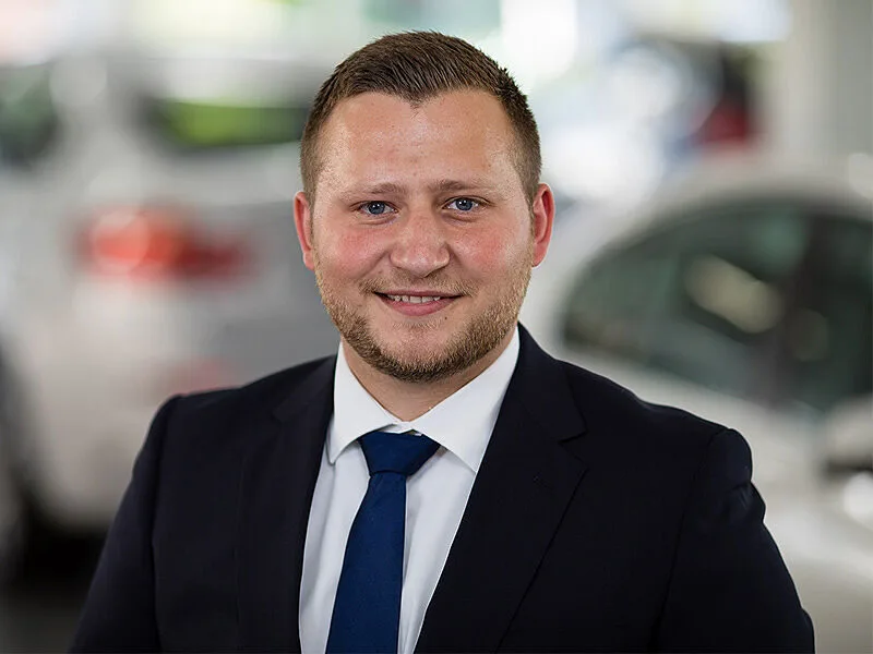 Kaltenbach Gruppe – Ein Mann im Anzug und mit blauer Krawatte lächelt in die Kamera. Er steht in einem scheinbaren Ausstellungsraum von Kaltenbach Remscheid mit unscharfen Fahrzeugen im Hintergrund. – BMW, MINI, KIA, BMW Motorrad & Honda Motorrad Partner in NRW