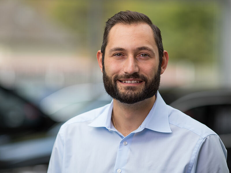 Kaltenbach Gruppe – Ein Mann mit kurzen dunklen Haaren und Bart, der ein hellblaues Button-up-Hemd trägt, lächelt in die Kamera. Im unscharfen Hintergrund sind Autos und Grünflächen zu sehen, die an die lebendige Atmosphäre von Kaltenbach Werdohl erinnern. – BMW, MINI, KIA, BMW Motorrad & Honda Motorrad Partner in NRW