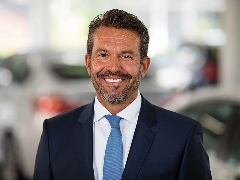 Kaltenbach Gruppe – Ein Mann im dunklen Anzug und mit blauer Krawatte lächelt in die Kamera. Er steht in einem Innenraum in Kaltenbach-Remscheid, mit unscharfen Autos und heller Beleuchtung im Hintergrund. – BMW, MINI, KIA, BMW Motorrad & Honda Motorrad Partner in NRW
