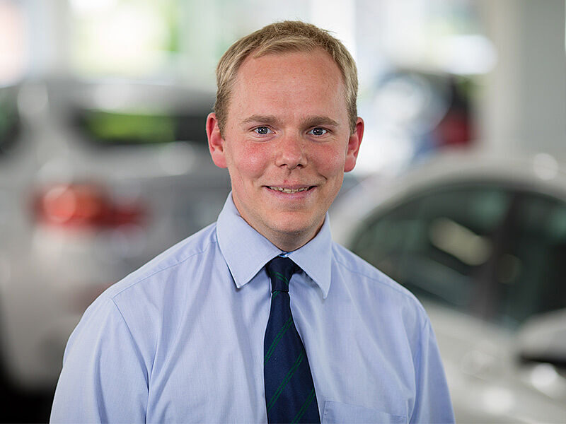 Kaltenbach Gruppe – Ein lächelnder Mann mit hellblauem Hemd und dunkler Krawatte steht in einem Innenraum vor unscharfen Autos, möglicherweise im Autohaus oder Ausstellungsraum von Kaltenbach Remscheid. – BMW, MINI, KIA, BMW Motorrad & Honda Motorrad Partner in NRW