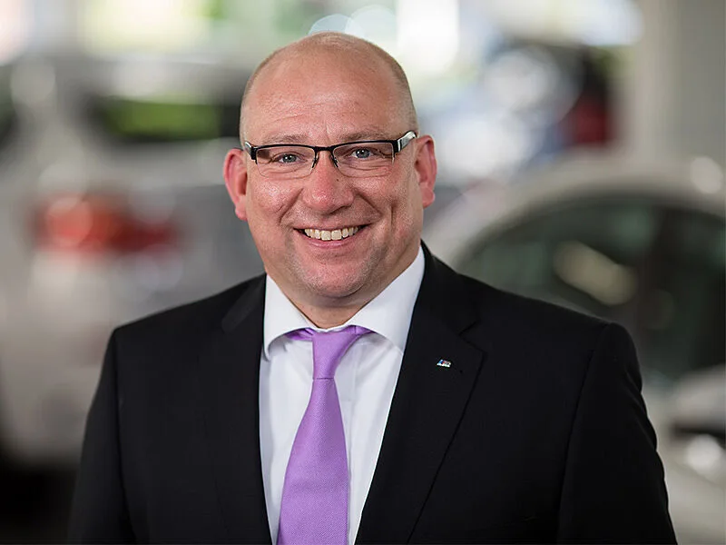 Kaltenbach Gruppe – Ein lächelnder Mann im Anzug, mit hellvioletter Krawatte und Brille steht in einem Haus in Kaltenbach-Remscheid, mit unscharfen Autos im Hintergrund. – BMW, MINI, KIA, BMW Motorrad & Honda Motorrad Partner in NRW