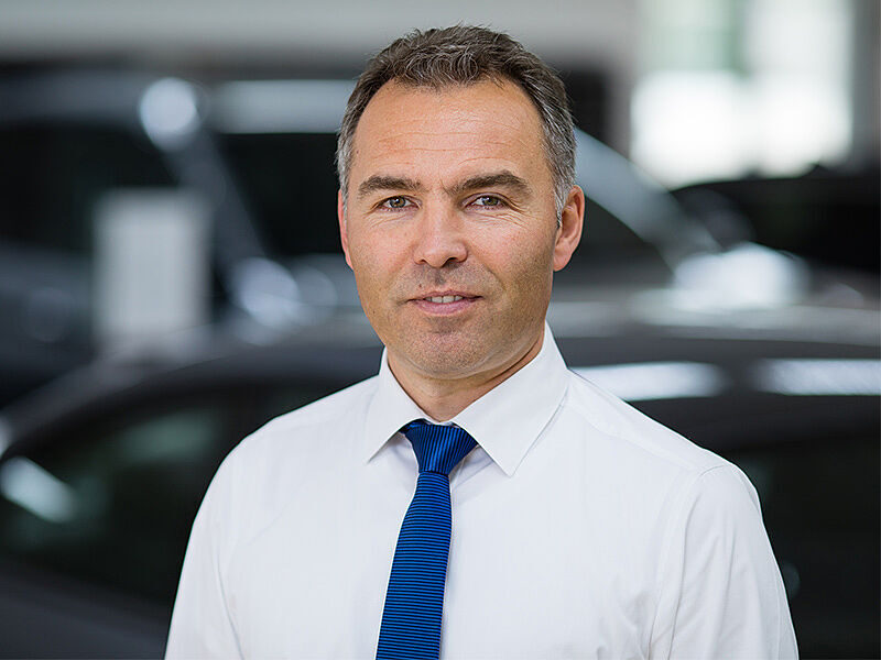 Kaltenbach Gruppe – Ein Mann mit weißem Hemd und blauer Krawatte steht in einer Halle in Kaltenbach Waldbröl vor einem unscharfen Hintergrund mit Autos, blickt direkt in die Kamera und lächelt leicht. – BMW, MINI, KIA, BMW Motorrad & Honda Motorrad Partner in NRW