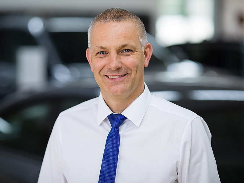 Kaltenbach Gruppe – Ein Mann mit weißem Hemd und blauer Krawatte lächelt in die Kamera, während er in einem Haus in Kaltenbach Waldbröl steht, mit unscharfen Autos im Hintergrund. – BMW, MINI, KIA, BMW Motorrad & Honda Motorrad Partner in NRW