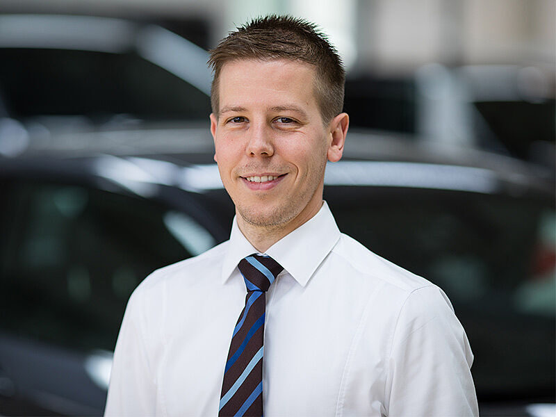 Kaltenbach Gruppe – Ein Mann mit kurzen braunen Haaren, weißem Hemd und gestreifter Krawatte lächelt, während er in einem Haus in Kaltenbach Waldbröl vor einem verschwommenen Hintergrund mit Autos steht. – BMW, MINI, KIA, BMW Motorrad & Honda Motorrad Partner in NRW