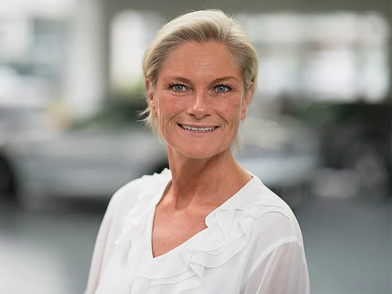 Kaltenbach Gruppe – Eine Frau mit kurzen blonden Haaren und weißer Bluse lächelt in die Kamera. Sie steht in der hell erleuchteten Kaltenbach-Zentrale, der Hintergrund ist unscharf. – BMW, MINI, KIA, BMW Motorrad & Honda Motorrad Partner in NRW