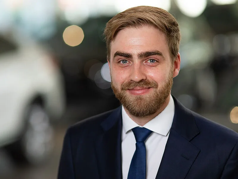 Kaltenbach Gruppe – Ein Mann mit hellbraunem Haar und Bart, der einen marineblauen Anzug, ein weißes Hemd und eine blaue Krawatte trägt, lächelt vor einem unscharfen Hintergrund in die Kamera - ein professioneller Moment in Kaltenbach-Wiehl. – BMW, MINI, KIA, BMW Motorrad & Honda Motorrad Partner in NRW