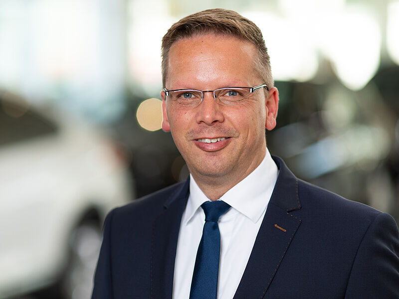 Kaltenbach Gruppe – Ein Mann in einem marineblauen Anzug, weißem Hemd und blauer Krawatte lächelt in die Kamera. Er hat kurzes hellbraunes Haar, eine Brille und steht in einem Haus in Kaltenbach-Wiehl, mit unscharfen Autos im Hintergrund. – BMW, MINI, KIA, BMW Motorrad & Honda Motorrad Partner in NRW