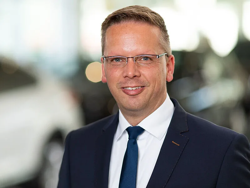 Kaltenbach Gruppe – Ein Mann in einem marineblauen Anzug, weißem Hemd und blauer Krawatte lächelt in die Kamera. Er hat kurzes hellbraunes Haar, eine Brille und steht in einem Haus in Kaltenbach-Wiehl, mit unscharfen Autos im Hintergrund. – BMW, MINI, KIA, BMW Motorrad & Honda Motorrad Partner in NRW