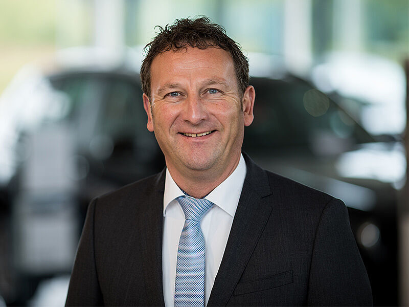 Kaltenbach Gruppe – Ein Mann in einem dunklen Anzug, weißem Hemd und hellblauer Krawatte lächelt in die Kamera. Er hat kurzes lockiges Haar und steht in einer unscharfen Innenraumkulisse mit Autos im Hintergrund in Kaltenbach Olpe. – BMW, MINI, KIA, BMW Motorrad & Honda Motorrad Partner in NRW