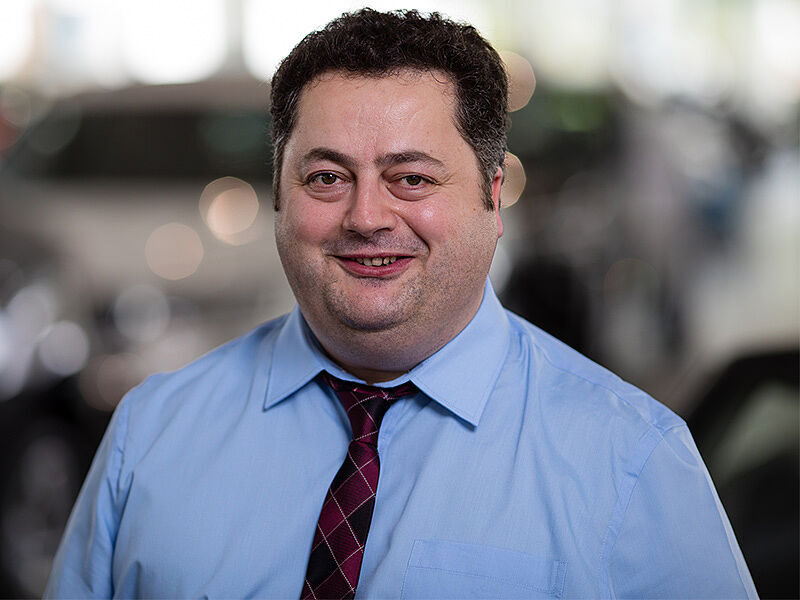 Kaltenbach Gruppe – Ein Mann mit kurzen dunklen Haaren, der ein hellblaues Hemd und eine rot gemusterte Krawatte trägt, lächelt, während er in einem Haus in Kaltenbach-Wiehl steht, mit unscharfen Autos im Hintergrund. – BMW, MINI, KIA, BMW Motorrad & Honda Motorrad Partner in NRW