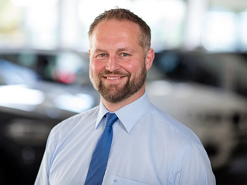 Kaltenbach Gruppe – Ein lächelnder Mann mit Bart und kurzen Haaren, der ein hellblaues Hemd und eine blaue Krawatte trägt, steht in einem Haus in Kaltenbach Wipperfürth, mit unscharfen Autos im Hintergrund. – BMW, MINI, KIA, BMW Motorrad & Honda Motorrad Partner in NRW