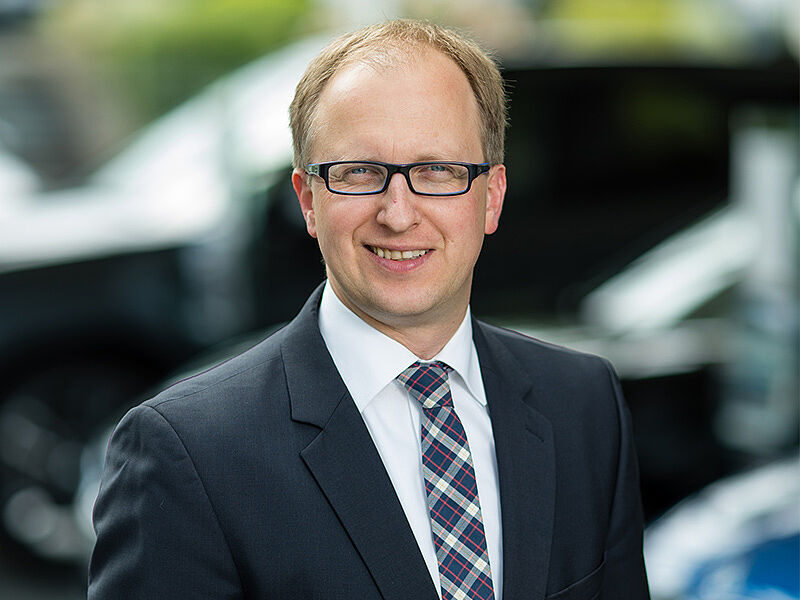 Kaltenbach Gruppe – Ein Mann mit kurzen hellbraunen Haaren und Brille, der einen dunklen Anzug, ein weißes Hemd und eine karierte Krawatte trägt, steht im Freien in Kaltenbach Waldbröl und lächelt in die Kamera. Der Hintergrund ist unscharf mit Andeutungen von Autos und Grünflächen. – BMW, MINI, KIA, BMW Motorrad & Honda Motorrad Partner in NRW