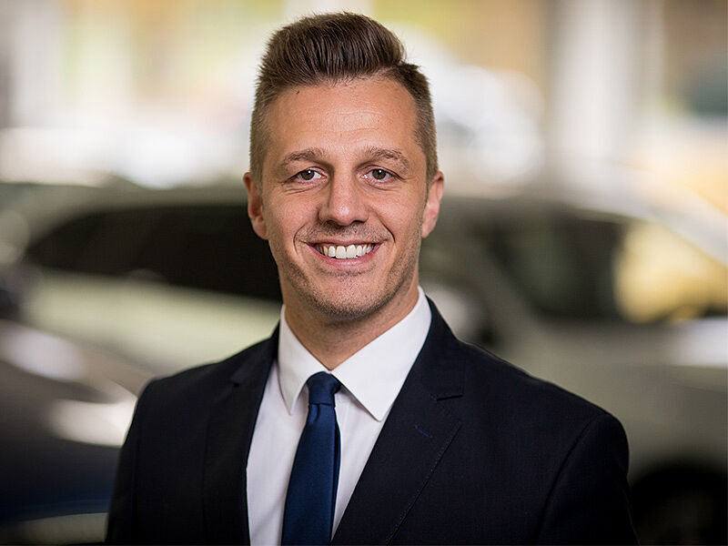 Kaltenbach Gruppe – Ein Mann in einem dunklen Anzug, weißem Hemd und blauer Krawatte lächelt in die Kamera. Er hat kurzes, gestyltes Haar und steht in einem Innenraum in Kaltenbach Overath, mit unscharfen Autos im Hintergrund. – BMW, MINI, KIA, BMW Motorrad & Honda Motorrad Partner in NRW