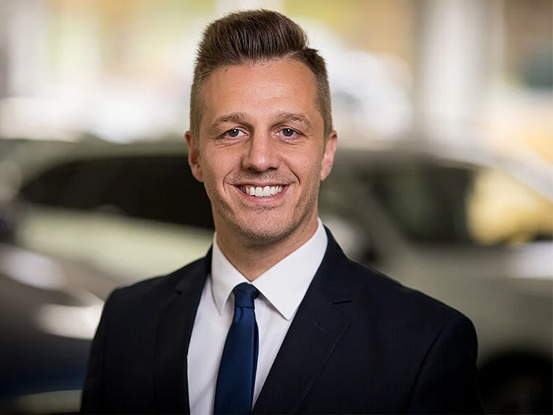 Kaltenbach Gruppe – Ein Mann in einem dunklen Anzug, weißem Hemd und blauer Krawatte lächelt in die Kamera. Er hat kurzes, gestyltes Haar und steht in einem Innenraum in Kaltenbach Overath, mit unscharfen Autos im Hintergrund. – BMW, MINI, KIA, BMW Motorrad & Honda Motorrad Partner in NRW
