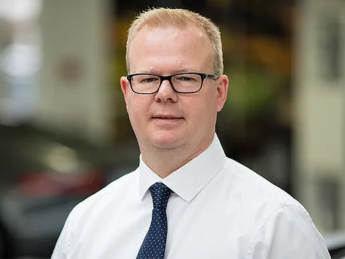Kaltenbach Gruppe – Ein Mann mit kurzen blonden Haaren und Brille, der ein weißes Hemd und eine marineblaue Krawatte trägt, steht in einem Haus mit unscharfem Hintergrund, möglicherweise in Kaltenbach-Meinerzhagen. – BMW, MINI, KIA, BMW Motorrad & Honda Motorrad Partner in NRW