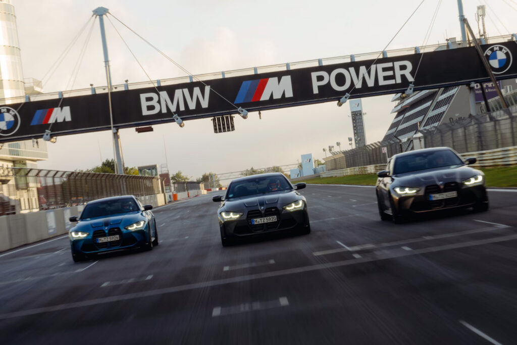 Kaltenbach Gruppe – Drei BMW Fahrzeuge fahren nebeneinander auf einer Rennstrecke unter einem großen BMW M Power Schild, mit eingeschalteten Scheinwerfern und unscharfen Bewegungen, die eine hohe Geschwindigkeit suggerieren. Die Strecke wird von Absperrungen und modernen Gebäuden flankiert. – BMW, MINI, KIA, BMW Motorrad & Honda Motorrad Partner in NRW