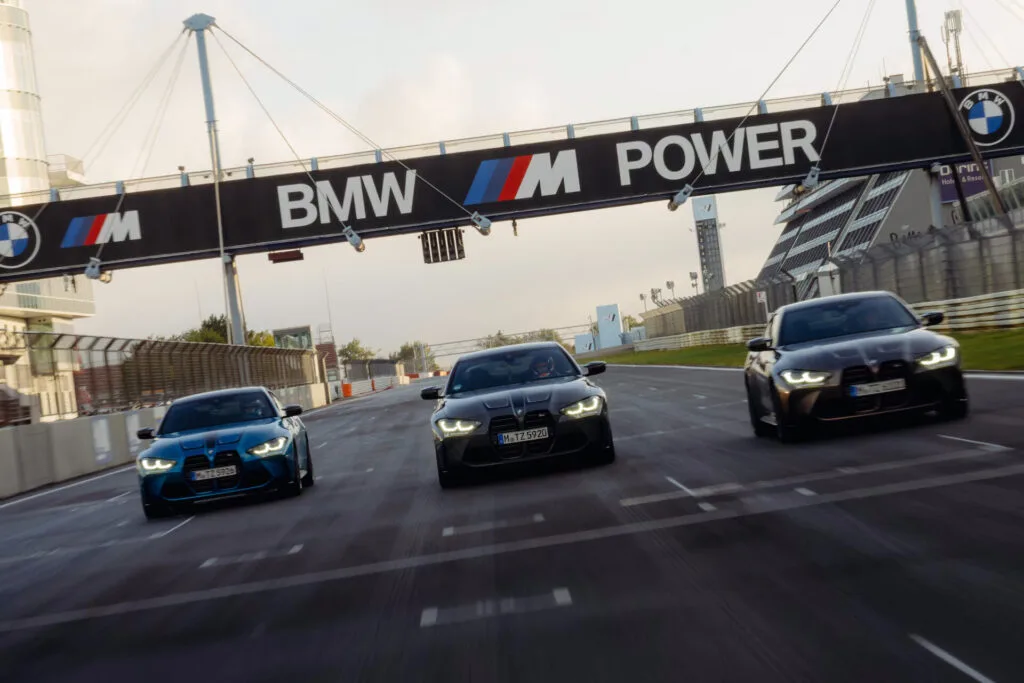 Kaltenbach Gruppe – Drei BMW Fahrzeuge fahren nebeneinander auf einer Rennstrecke unter einem großen BMW M Power Schild, mit eingeschalteten Scheinwerfern und unscharfen Bewegungen, die eine hohe Geschwindigkeit suggerieren. Die Strecke wird von Absperrungen und modernen Gebäuden flankiert. – BMW, MINI, KIA, BMW Motorrad & Honda Motorrad Partner in NRW
