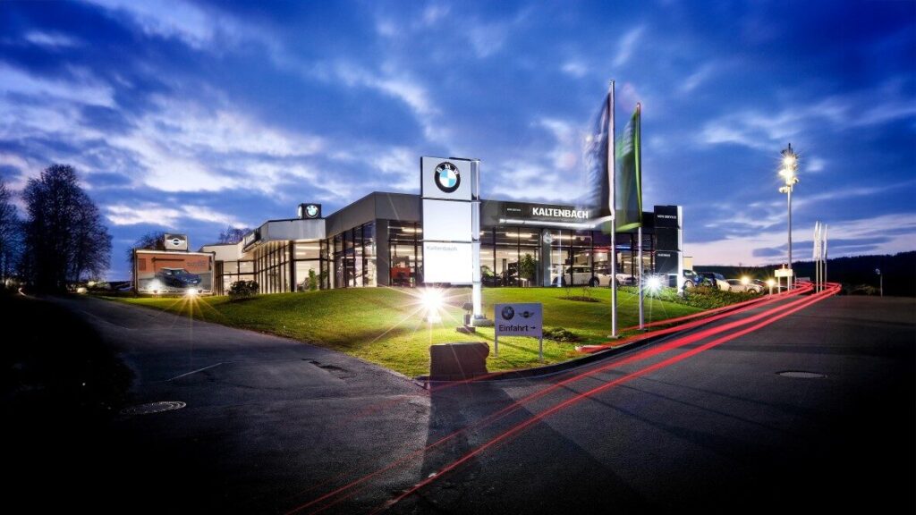 Kaltenbach Gruppe – Ein modernes BMW-Händlergebäude ist in der Dämmerung mit hellen Lichtern und sichtbaren BMW-Schildern beleuchtet. Davor sind Fahnen und ein gepflegter Rasen zu sehen, und rote Streifen von Autoscheinwerfern ziehen sich über die Straße im Vordergrund. – BMW, MINI, KIA, BMW Motorrad & Honda Motorrad Partner in NRW