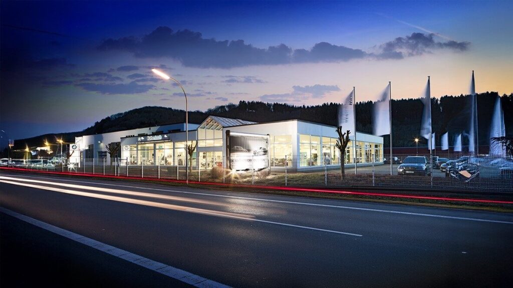 Kaltenbach Gruppe – Ein modernes Autohaus mit großen Glasfenstern ist in der Abenddämmerung beleuchtet, und ein Lastwagen steht am Eingang. Mehrere weiße Fahnen säumen den umzäunten Bereich, und auf der Straße davor sind die Lichtspuren der vorbeifahrenden Autos zu sehen. – BMW, MINI, KIA, BMW Motorrad & Honda Motorrad Partner in NRW