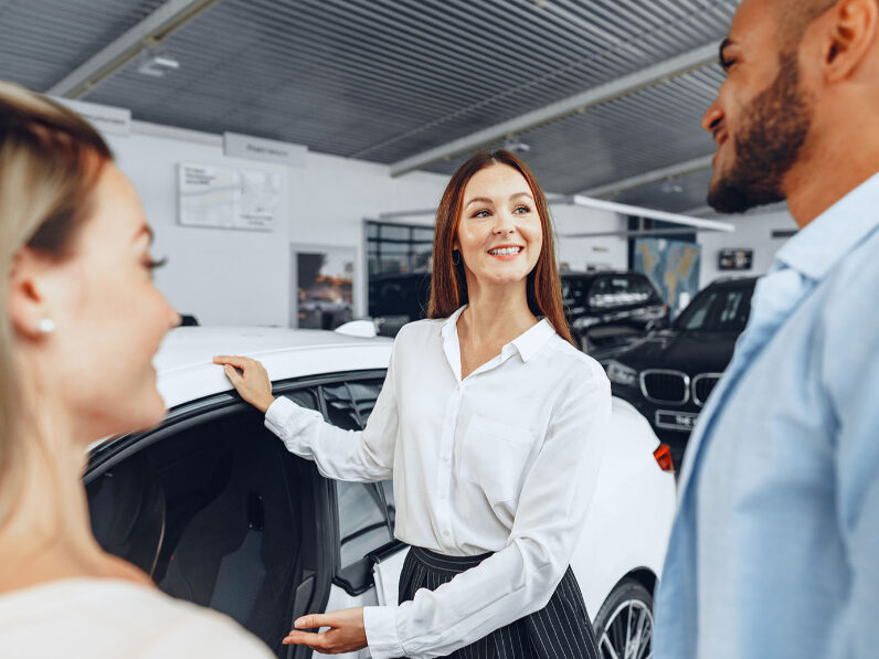 Kaltenbach Gruppe – Eine Frau in Geschäftskleidung zeigt einem lächelnden Paar in einem Autohaus ein Auto, was darauf hindeutet, dass sie über das Fahrzeug diskutieren oder es in Betracht ziehen. Im Hintergrund sind andere Autos zu sehen. – BMW, MINI, KIA, BMW Motorrad & Honda Motorrad Partner in NRW