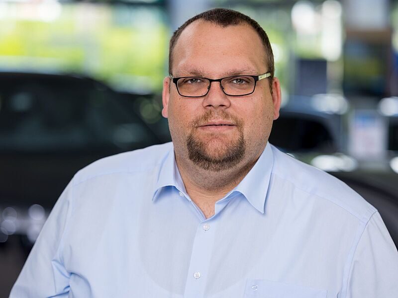 Kaltenbach Gruppe – Ein Mann mit kurzen braunen Haaren, Brille und gestutztem Bart, der ein hellblaues Button-up-Hemd trägt, steht in einem Innenraum in Kaltenbach-Wiehl mit unscharfen Autos und Grünflächen im Hintergrund. – BMW, MINI, KIA, BMW Motorrad & Honda Motorrad Partner in NRW