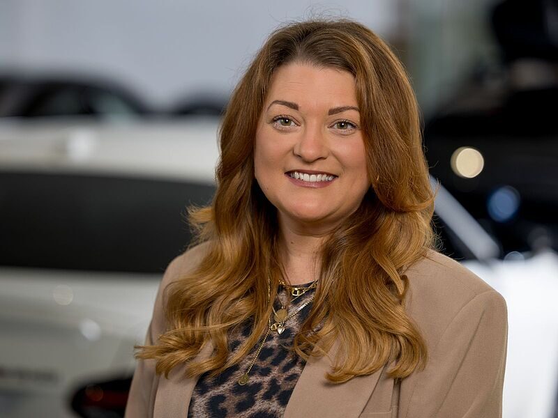 Kaltenbach Gruppe – Eine Frau mit langen, gewellten hellbraunen Haaren lächelt in die Kamera. Sie trägt einen beigen Blazer über einem Oberteil mit Leopardenmuster und steht in einem Innenraum der Kaltenbach-Zentrale mit unscharfen Autos im Hintergrund. – BMW, MINI, KIA, BMW Motorrad & Honda Motorrad Partner in NRW