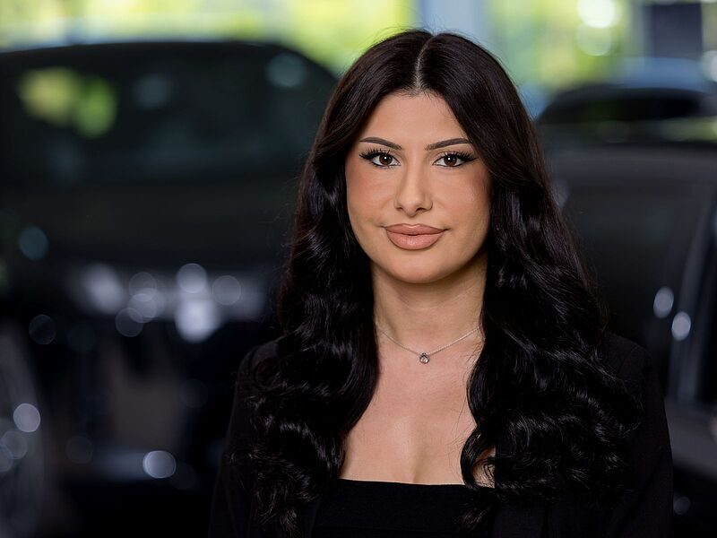 Kaltenbach Gruppe – Eine Frau mit langen, gewellten schwarzen Haaren und natürlichem Make-up lächelt leicht, während sie in einem Haus in Kaltenbach-Wiehl vor verschwommenen dunklen Autos steht. Sie trägt ein schwarzes Oberteil und eine zarte Halskette. – BMW, MINI, KIA, BMW Motorrad & Honda Motorrad Partner in NRW