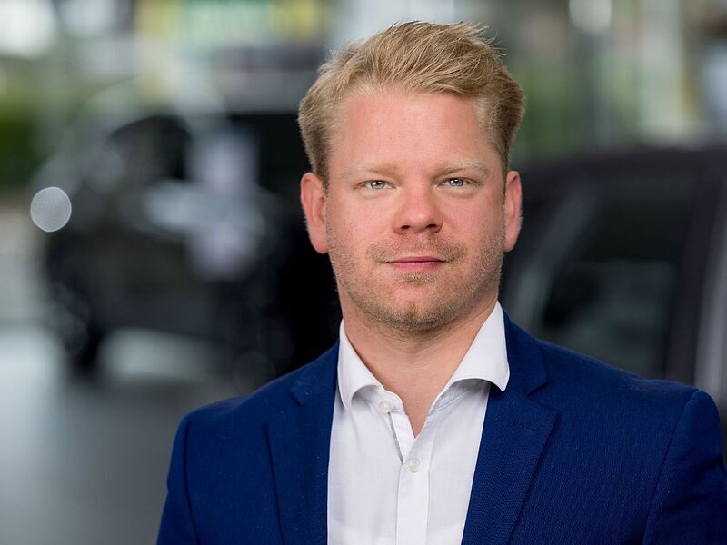 Kaltenbach Gruppe – Ein Mann mit kurzem blondem Haar und gestutztem Bart, der eine blaue Anzugsjacke und ein weißes Hemd trägt, steht in einem Innenraum mit unscharfem Hintergrund, möglicherweise in einem Büro oder Ausstellungsraum von Kaltenbach Overath. – BMW, MINI, KIA, BMW Motorrad & Honda Motorrad Partner in NRW