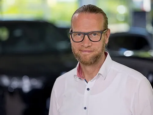 Kaltenbach Gruppe – Ein Mann mit heller Haut, Brille und Bart lächelt sanft in einem weißen Button-up-Hemd. Der unscharfe Hintergrund suggeriert einen Innenraum in Kaltenbach Meinerzhagen, hinter ihm sind Autos zu sehen. – BMW, MINI, KIA, BMW Motorrad & Honda Motorrad Partner in NRW