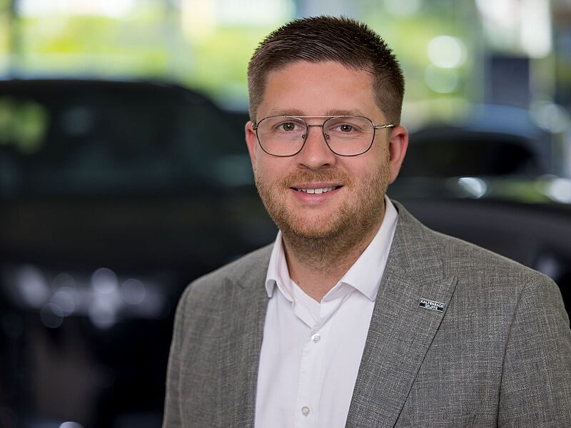 Kaltenbach Gruppe – Ein Mann mit kurzen braunen Haaren, Brille und Bart, der einen grauen Blazer und ein weißes Hemd trägt, lächelt, während er in einem Innenraum der Kaltenbach-Zentrale steht, mit verschwommenen dunklen Autos im Hintergrund. – BMW, MINI, KIA, BMW Motorrad & Honda Motorrad Partner in NRW