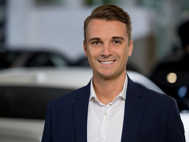 Kaltenbach Gruppe – Ein lächelnder junger Mann in einem marineblauen Blazer und weißem Hemd steht in der Kaltenbach Zentrale, mit verschwommenen Autos und einem hellen Hintergrund hinter ihm. – BMW, MINI, KIA, BMW Motorrad & Honda Motorrad Partner in NRW