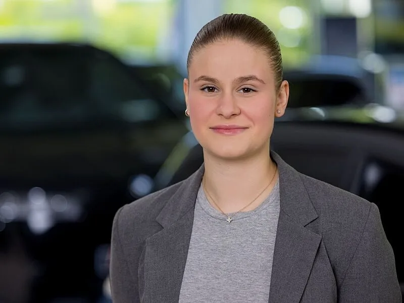 Kaltenbach Gruppe – Eine junge Frau mit heller Haut und zurückgebundenen braunen Haaren, die einen grauen Blazer und ein graues Oberteil trägt, steht in einem Innenraum in Kaltenbach Wermelskirchen mit unscharfen Autos im Hintergrund. Sie blickt in die Kamera und lächelt leicht. – BMW, MINI, KIA, BMW Motorrad & Honda Motorrad Partner in NRW