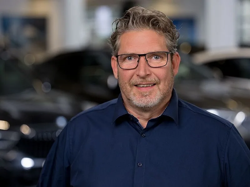 Kaltenbach Gruppe – Ein Mann mittleren Alters mit gewelltem grauem Haar, Brille und Bart lächelt und trägt ein marineblaues Hemd. Er steht in einem Innenraum in Kaltenbach Remscheid, mit verschwommenen Autos und einem hellen Hintergrund hinter ihm. – BMW, MINI, KIA, BMW Motorrad & Honda Motorrad Partner in NRW