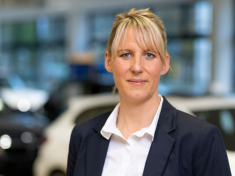 Kaltenbach Gruppe – Eine Frau mit blonden Haaren, weißem Hemd und dunklem Blazer, steht in einer Halle in Kaltenbach Overath vor unscharfen Autos und schaut mit neutralem Blick in die Kamera. – BMW, MINI, KIA, BMW Motorrad & Honda Motorrad Partner in NRW