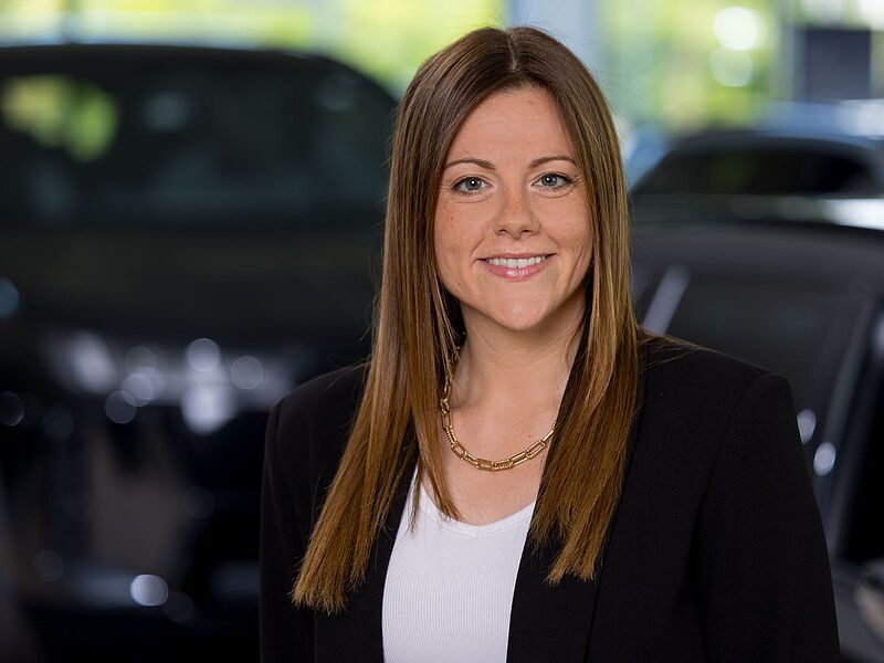 Kaltenbach Gruppe – Eine Frau mit glattem braunem Haar, die einen schwarzen Blazer, ein weißes Oberteil und eine goldene Halskette trägt, lächelt in einer Halle in Kaltenbach Waldbröl, mit verschwommenen Autos im Hintergrund. – BMW, MINI, KIA, BMW Motorrad & Honda Motorrad Partner in NRW