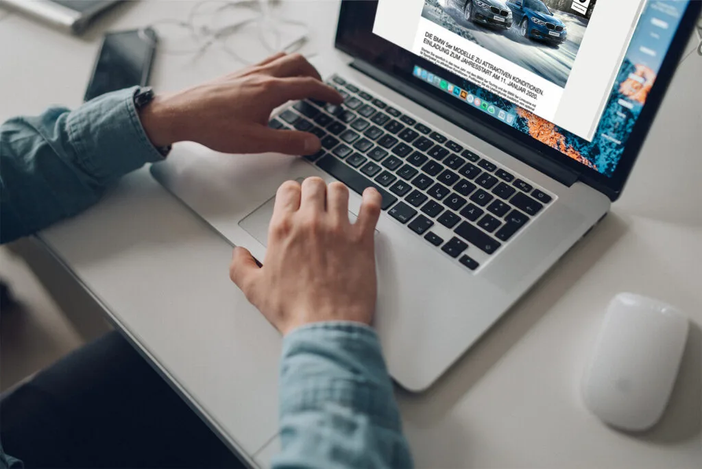 Kaltenbach Gruppe – Eine Person tippt auf einem silbernen Laptop an einem weißen Schreibtisch, in dessen Nähe sich ein Smartphone, Kopfhörer und eine weiße drahtlose Maus befinden. Auf dem Laptop-Bildschirm wird eine Website mit Autos und Text angezeigt. – BMW, MINI, KIA, BMW Motorrad & Honda Motorrad Partner in NRW