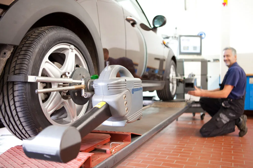 Kaltenbach Gruppe – Ein Mechaniker kniet neben einem Auto mit einem Achsmessgerät am Vorderreifen in einer Autowerkstatt und konzentriert sich auf die Reparatur nach einem Unfall. Ein Computer im Hintergrund zeigt präzise Daten zur Achsvermessung an. – BMW, MINI, KIA, BMW Motorrad & Honda Motorrad Partner in NRW