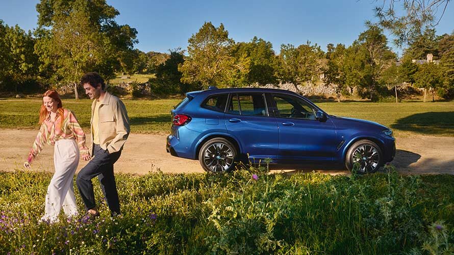 Kaltenbach Gruppe – Ein Paar spaziert lächelnd Hand in Hand über eine Wiese mit Wildblumen, während ihr blauer Geländewagen - mit flexiblen Servicepaketen - an einem sonnigen Tag auf einem Feldweg, umgeben von grünen Bäumen, steht. – BMW, MINI, KIA, BMW Motorrad & Honda Motorrad Partner in NRW