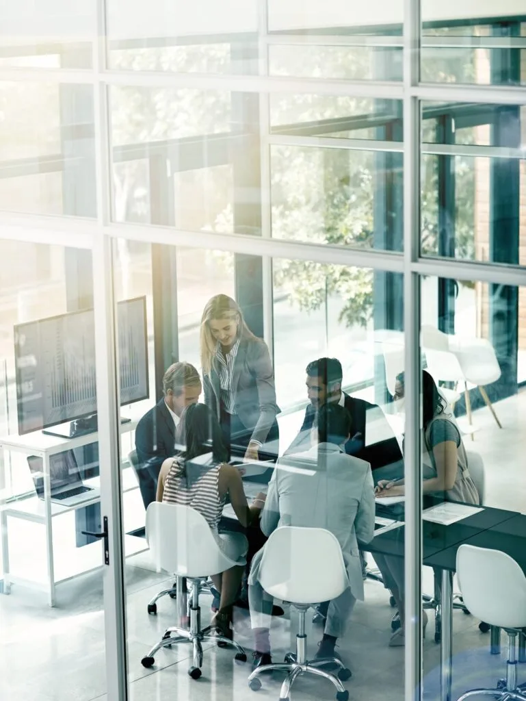 Kaltenbach Gruppe – Eine Gruppe von sechs Businesskunden sitzt in einem modernen Büro mit Glaswänden an einem Tisch, während im Hintergrund Diagramme auf einem Bildschirm angezeigt werden. Die Atmosphäre wirkt kollegial und professionell. – BMW, MINI, KIA, BMW Motorrad & Honda Motorrad Partner in NRW