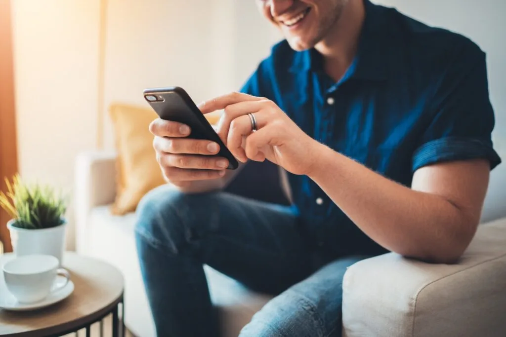 Kaltenbach Gruppe – Ein lächelnder Mann in einem blauen Hemd sitzt auf einer Couch und benutzt ein Smartphone, vielleicht verwaltet er Businesskunden. Neben ihm steht ein kleiner Tisch mit einer Topfpflanze und einer Tasse. Warmes Sonnenlicht durchflutet den Raum. – BMW, MINI, KIA, BMW Motorrad & Honda Motorrad Partner in NRW