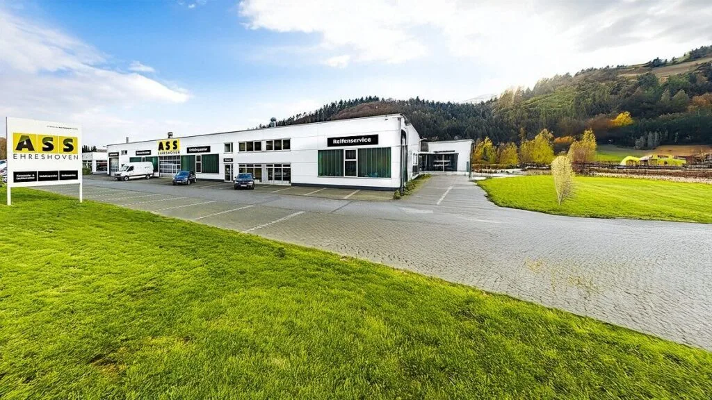 Kaltenbach Gruppe – Ein großes, modernes Gebäude mit "ASS"- und "Ehreshoven"-Schildern steht an einer grünen Wiese und Hügeln in Engelskirchen. Mehrere Autos sind vor dem Gebäude geparkt, im Hintergrund sind bei teilweise bewölktem Himmel die Berge zu sehen. – BMW, MINI, KIA, BMW Motorrad & Honda Motorrad Partner in NRW
