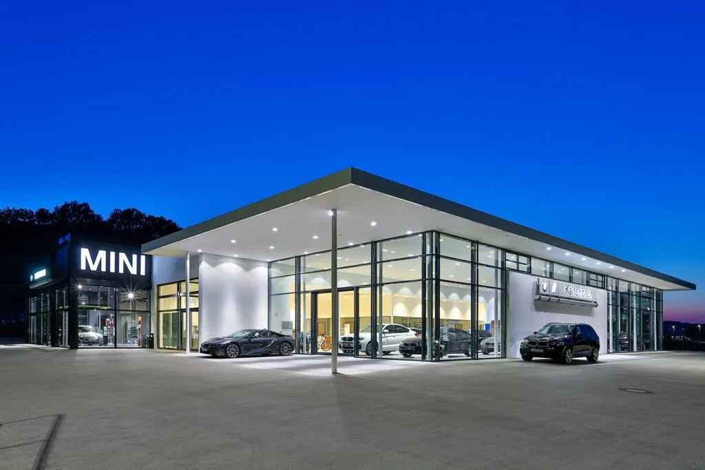 Kaltenbach Gruppe – Modernes Autohaus mit großen Glasfenstern, hell erleuchtetem Innenraum und mehreren Autos, die innen und außen zu sehen sind. Das minimalistische Gebäude zeigt ein MINI-Schild und den Namen Kaltenbach Olpe vor einem klaren Abendhimmel. – BMW, MINI, KIA, BMW Motorrad & Honda Motorrad Partner in NRW